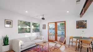 Un salon moderne avec un canapé blanc, une table basse, un tapis à motifs, une chaise en bois, une table à manger avec des chaises et de grandes fenêtres donnant sur un paysage verdoyant.