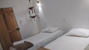 Una habitación con dos camas individuales, cada una con una almohada y una toalla. Se observa una puerta de madera y un colgador con luz.
