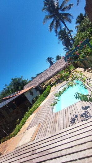 A wooden deck leading to a swimming pool with a thatched roof structure and palm trees in the background. - Pousada Al Mare (Cajueiro da Praia)