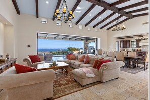 Uma sala de estar espaçosa com uma grande janela que oferece vista para o oceano, um espaço de jantar com mesa de madeira e uma cozinha com armários escuros.