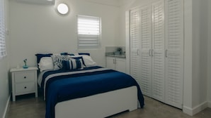 A bedroom with a bed, nightstand, wardrobe, and a window with blinds. - Timeless - Lovely beach cottage. Private pool, ocean view, beach access. R &R ! (Kingston)