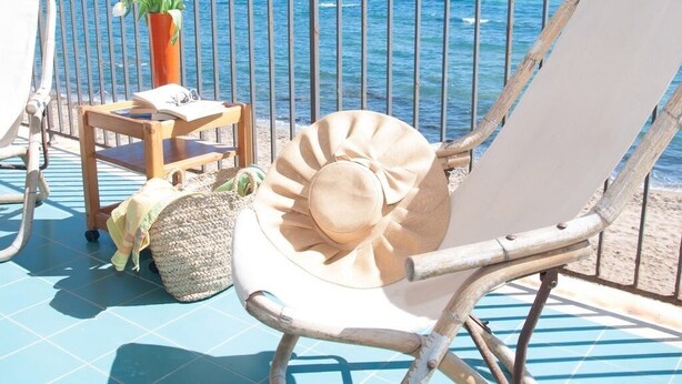 A sunlit outdoor seating area with a view of the sea, a wooden table with a vase of flowers, and a straw bag. - Apollo's Terraces-Beach apartments less than 20 m from the sea! Levant (Marinella)