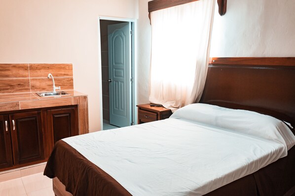 A bedroom with a wooden headboard, a single bed, a nightstand, a bedside table, and a window with curtains.