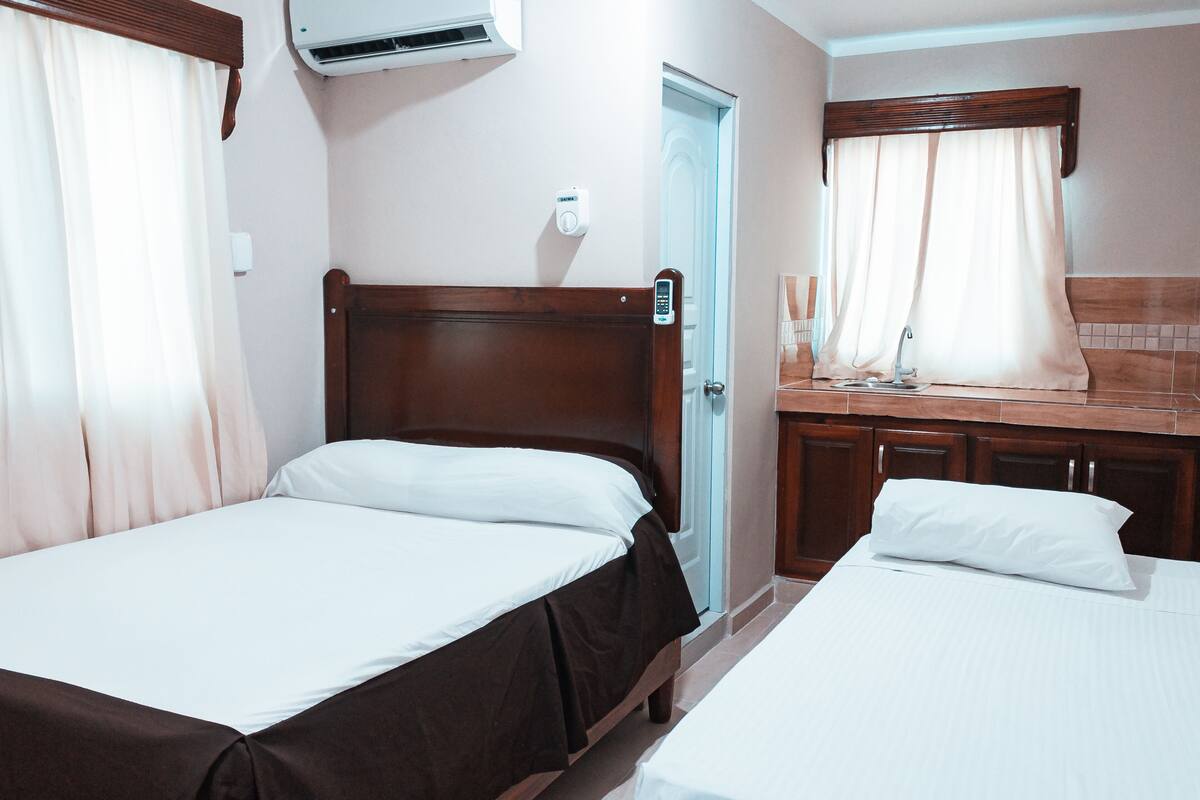 A hotel room with two single beds, a wooden headboard, a bathroom with a shower, and a small kitchenette.