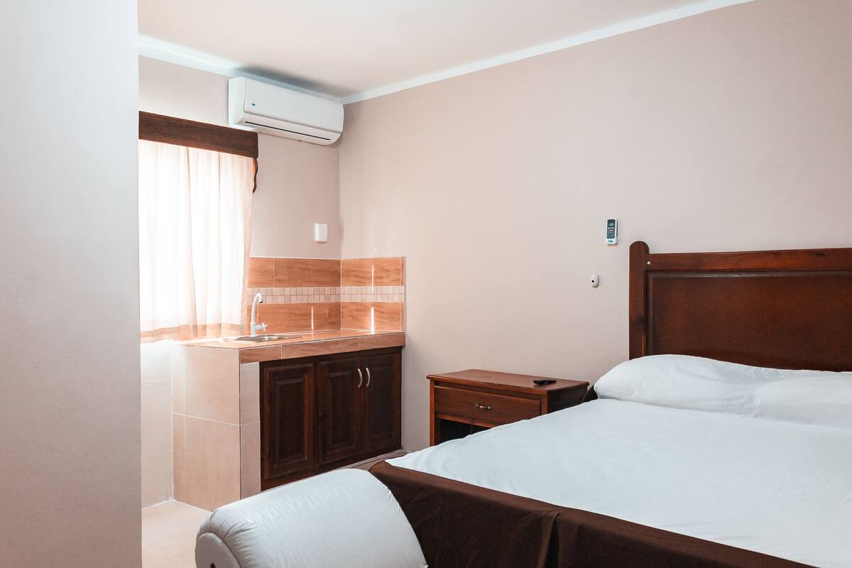 A hotel room with a bed, a wooden nightstand, a small table, a sink, and an air conditioning unit.
