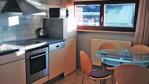 A compact kitchen with a stove, oven, and microwave. There's a glass table with chairs and a window with curtains. - Haus Rist - Apartment Panoramablick in Altstädten im Allgäu (Sonthofen)