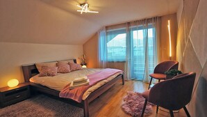A bedroom with a bed, bedside tables, a chair, a rug, and a window with curtains. - Haus Rist - Apartment Panoramablick in Altstädten im Allgäu (Sonthofen)