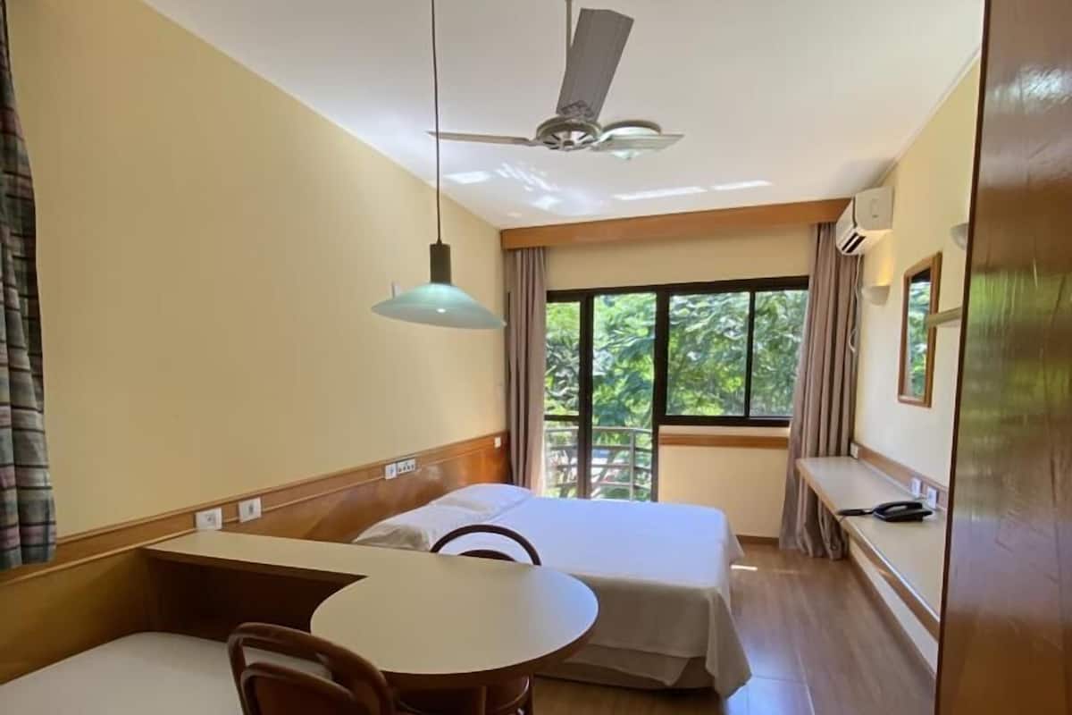 A room with a bed, a table with chairs, a ceiling fan, and a window with curtains.