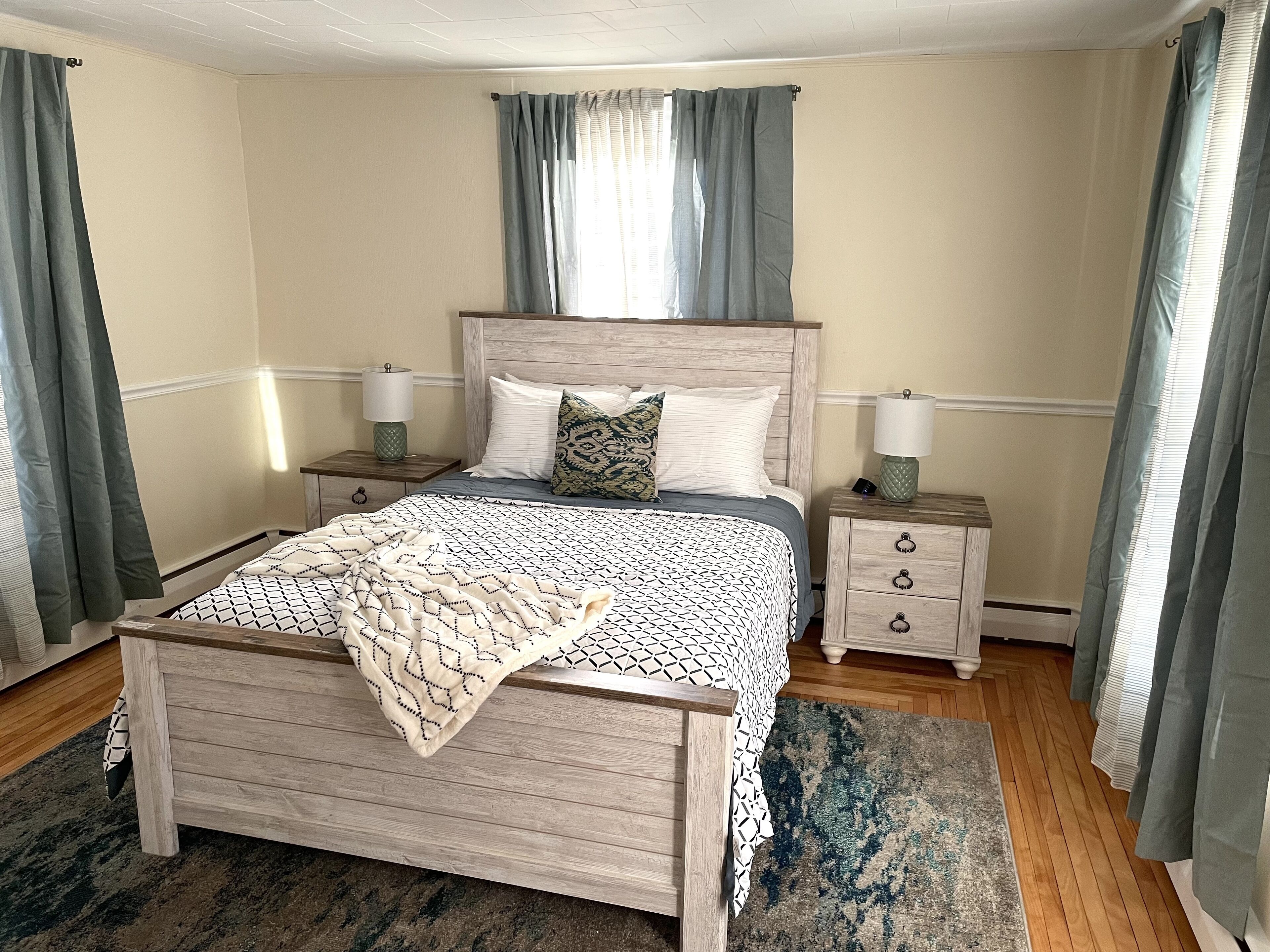 Une chambre à coucher avec un lit en bois, deux tables de chevet avec des lampes, une couvre-lit à motifs et un rideau rayé bleu et blanc.