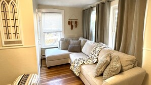 Ein gemütliches Wohnzimmer mit einem beigen Sofa, einer gemusterten Decke und einem Fenster mit Vorhängen.
