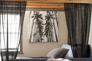 Une chambre à coucher avec un lit, une œuvre d’art représentant des palmiers accrochée au mur, des rideaux légers et un plafond en bois.