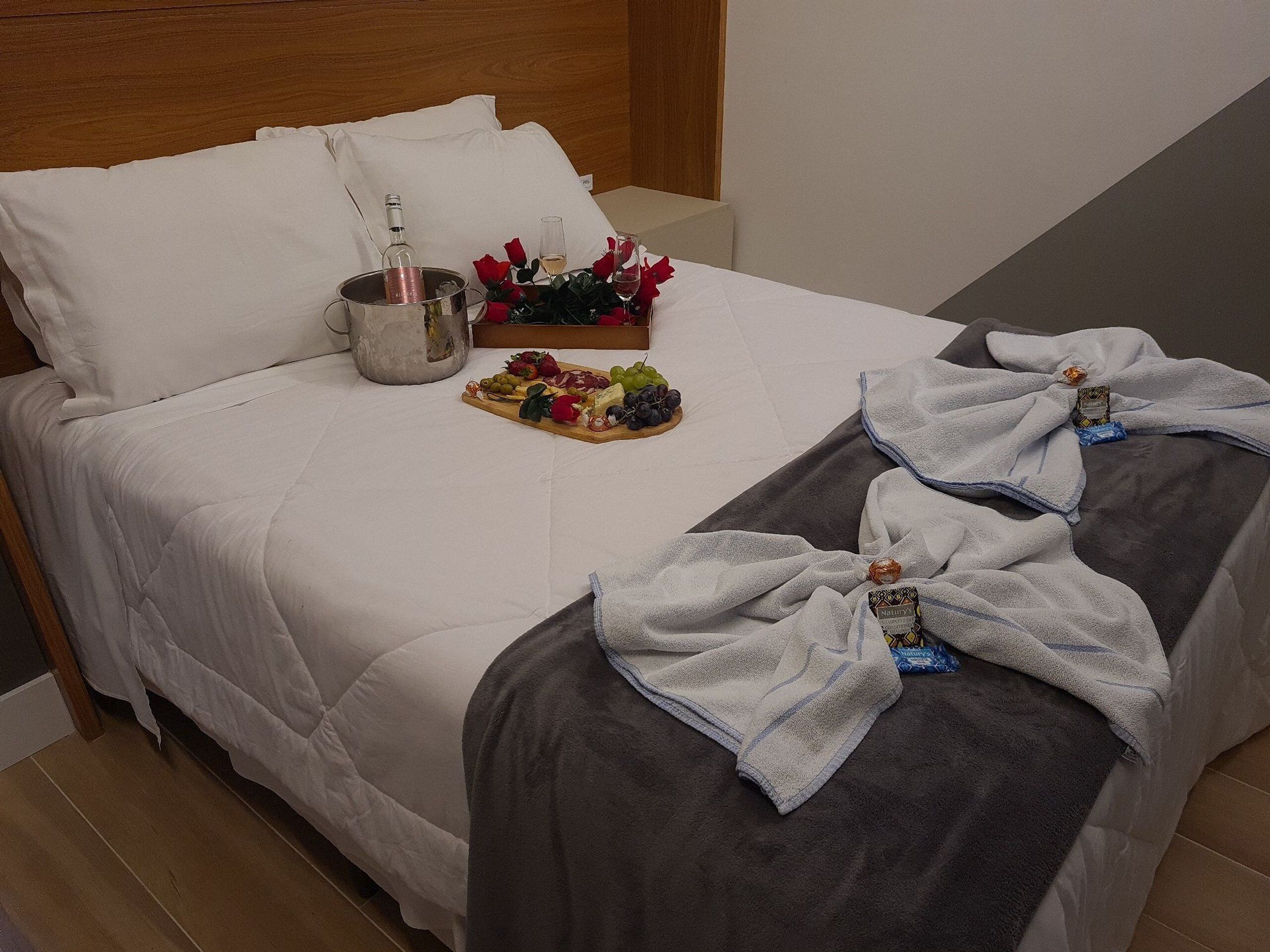 A neatly made bed with white linens, a fruit platter, a bottle of water, and a glass of wine on the bed.