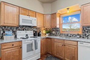A kitchen with wooden cabinets, a white stove, a microwave, a window with a view, and a stone backsplash. - Luxury Vacation Cabin on the White River (Cotter)