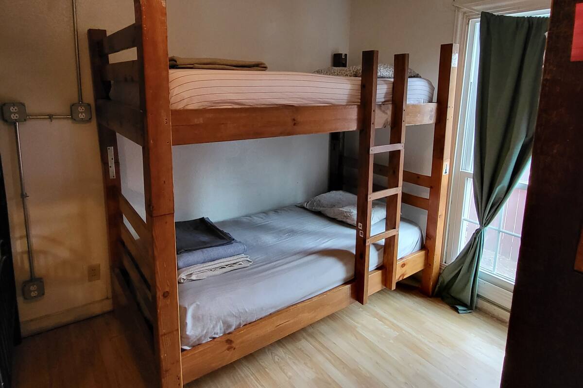 A bunk bed room with a wooden frame, a window with curtains, and a power outlet on the wall.