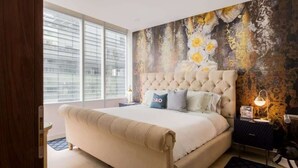 Een slaapkamer met een groot bed, een gestuwd hoofdeinde, een raam met jaloezieën, een muurschildering en een nachtkastje met een lamp.