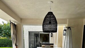 Uma área de refeições ao ar livre com uma mesa comprida e cadeiras, um lustre pendurado, e vista para um jardim e um edifício ao fundo.