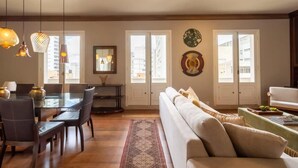 A spacious living area with a sofa, dining table, and chairs. There are two windows with white frames and a decorative wall piece. - Tiete Classic I Stunning 3 Bedroom in Jardins (São Paulo)