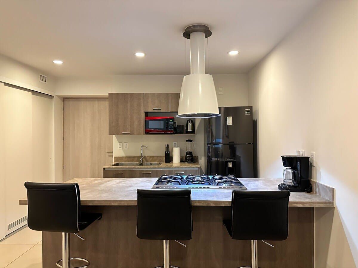 A modern kitchen with a central island, black bar stools, a stainless steel refrigerator, and a microwave.