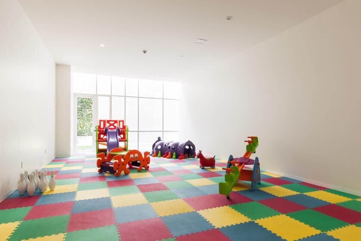 A brightly lit room with colorful interlocking foam mats on the floor, a play area with various toys and equipment, and a large window providing natural light.