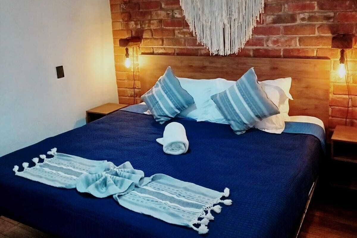 Une chambre avec un mur en briques, un lit avec une literie bleue, des oreillers blancs et une décoration murale en macramé.