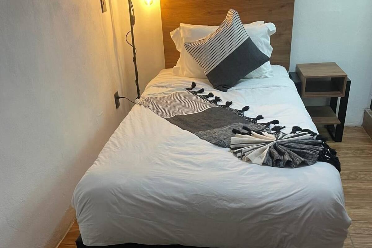 Un lit bien fait, avec du linge de lit blanc et un plaid rayé, une tête de lit en bois et une petite table de chevet en bois à côté.