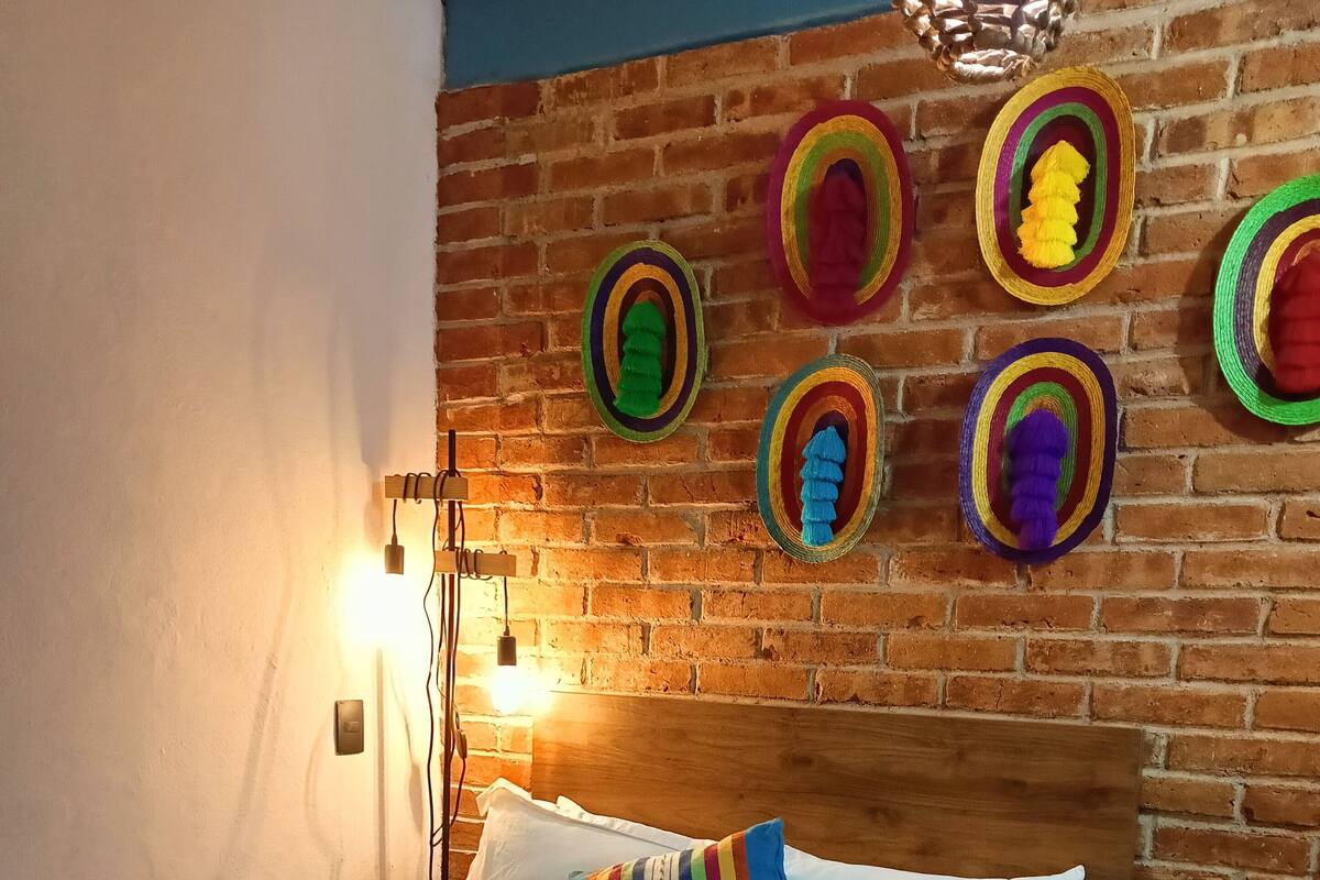 Un lit bien fait, avec du linge de lit blanc, un oreiller à rayures et une serviette enroulée. Une petite table de chevet en bois, sur laquelle se trouve une télécommande, est placée à côté du lit. La pièce est agrémentée d’un mur de briques sur lequel sont accrochés des paniers colorés en fibres tressées.