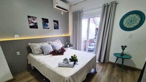 A hotel room with a bed, a small table with a vase, a wall-mounted air conditioner, and a window with curtains. - Duplex 02 Suites - Cond.Fechado 08 P (Gramado)