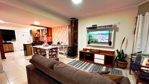 A living room with a sofa, a TV, a dining area, and a kitchen in the background. - Duplex 02 Suites - Cond.Fechado 08 P (Gramado)