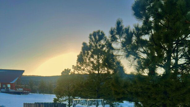 A wooden deck with two wine glasses on it, overlooking a snowy landscape with trees and a building in the background. - Three Bears Cabin (Angel Fire)