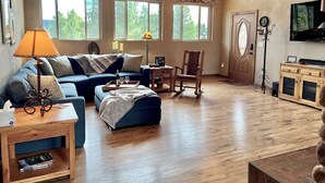 A spacious living room with a wooden ceiling, large windows, a blue sofa, a wooden rocking chair, and a wooden cabinet with a television. - Three Bears Cabin (Angel Fire)