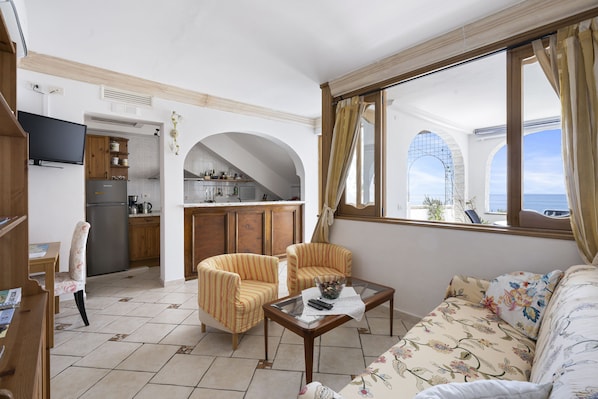 Una sala de estar con sofá, dos sillones, una mesa de centro y televisor. Se ve una cocina al fondo con refrigerador, estufa y alacenas.