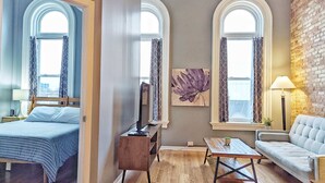 A hotel room with a bed, a desk, a chair, a sofa, and two windows with curtains. - Tanner Bldg Apt 7 - 2 bedroom 1 king & 1 queen (Downtown) (Bay City)