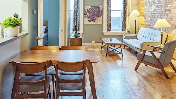 A dining area with a wooden table and chairs, a hanging light fixture, and a brick wall. - Tanner Bldg Apt 7 - 2 bedroom 1 king & 1 queen (Downtown) (Bay City)