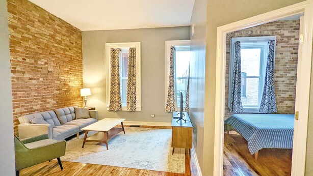 A cozy living room with a brick accent wall, a sofa, a coffee table, and a bed with striped bedding. - Tanner Bldg Apt 3 - 2 bedroom (Downtown) (Bay City)