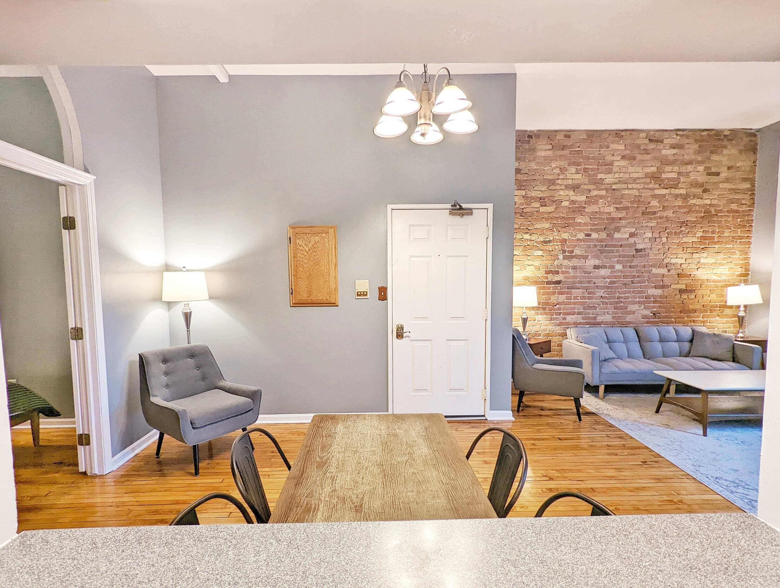 A dining area with a wooden table and chairs, a gray sofa, and a brick wall.