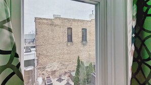 A window with a view of a brick building, a balcony with outdoor furniture, and a green patterned curtain. - Tanner Bldg Apt 3 - 2 bedroom (Downtown) (Bay City)