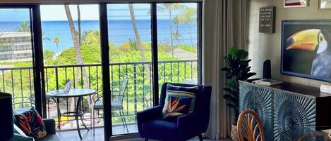 Ein Wohnzimmer mit einem großen Fenster und Blick auf den Ozean, einem Sofa, einem Sessel, einem kleinen Tisch und einem Fernseher.