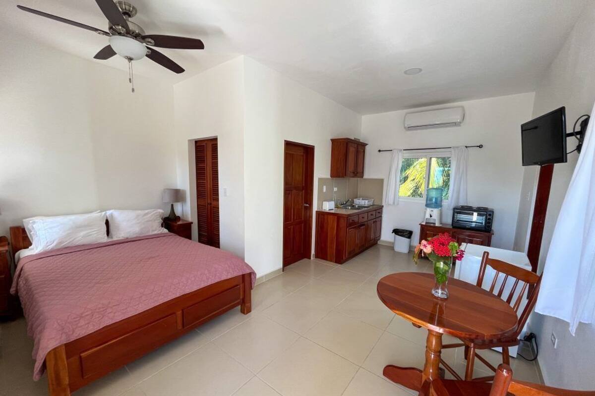 A bedroom with a bed, a dining table with chairs, a kitchen area, and a ceiling fan.