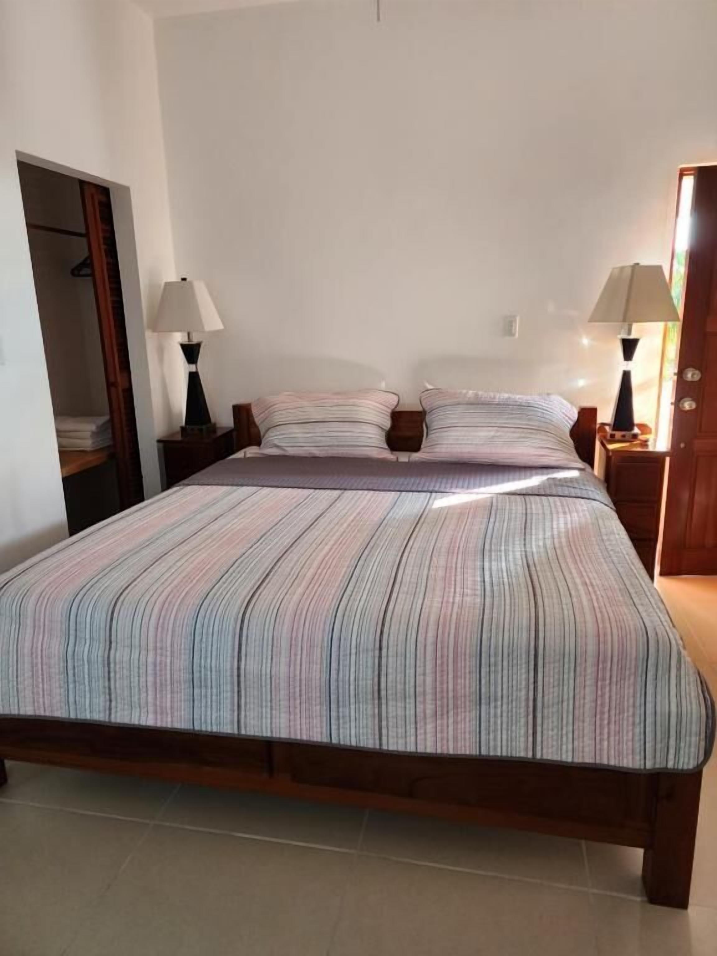 Un dormitorio con una cama grande, dos mesitas de noche con lámparas, un armario de madera y una puerta que da a otra habitación.