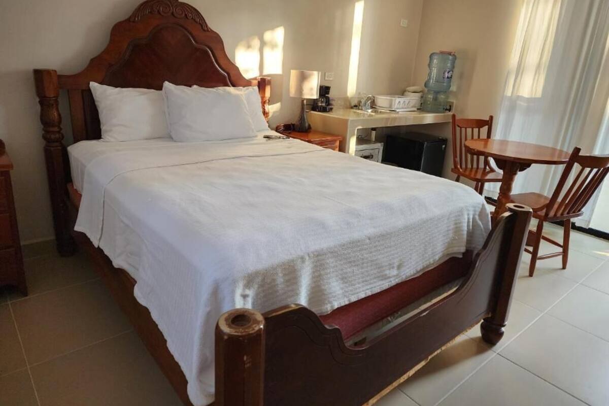 A neatly made bed with a wooden headboard, a bedside table with a lamp, a small desk with a chair, and a water dispenser.