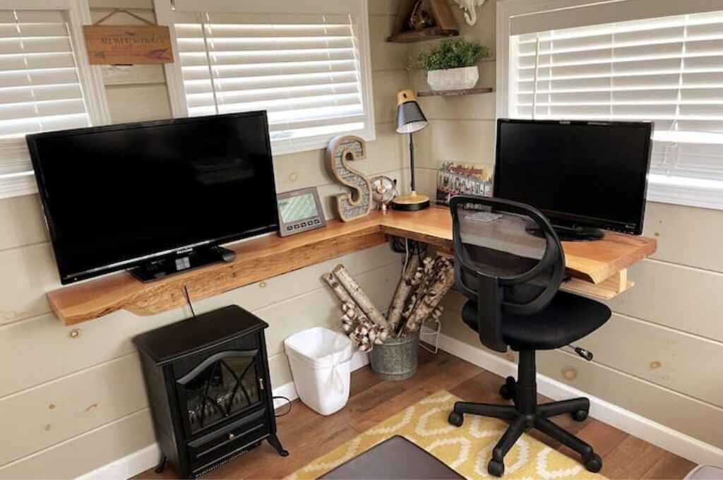 A home office with two monitors, a wooden desk, a black office chair, a fireplace, and a window with blinds.