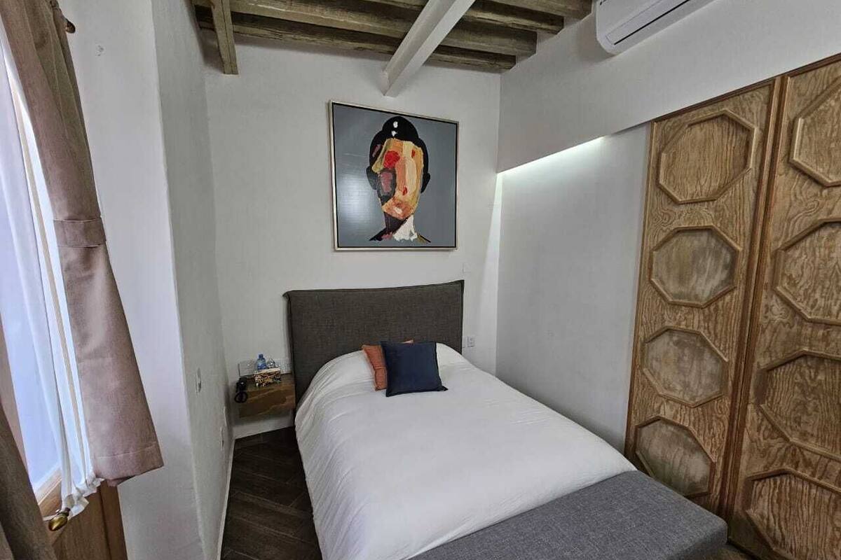 A bedroom with a bed, a headboard, a painting, a wardrobe, and a bedside table with a lamp.