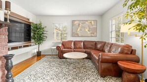 A living room with a brown leather sofa, a patterned rug, a fireplace, and a large window with blinds. - Hot tub, pool, bbq, game room near downtown (Ocala)