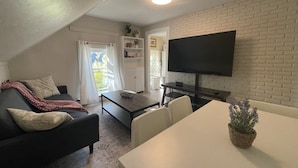 A compact living space with a sofa, a dining table, a TV mounted on a brick wall, and a window with curtains. - 2b apt loft Downtown Ipswich (Ipswich)