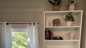 A room with a large window, white curtains, a bookshelf with books and decorative items, and a white cabinet with louvered doors. - 2b apt loft Downtown Ipswich (Ipswich)
