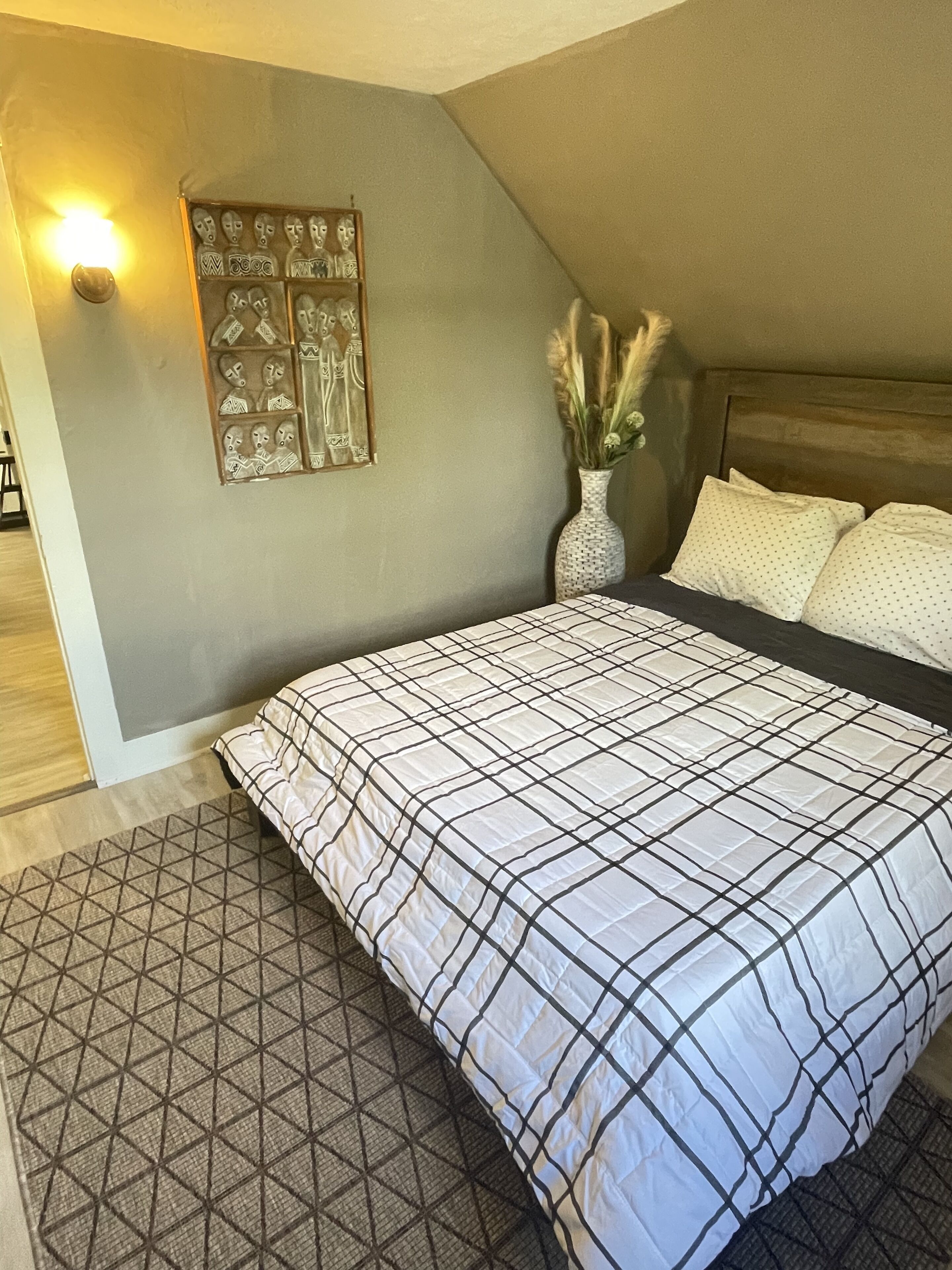 A bedroom with a large bed, a geometric-patterned rug, a wall-mounted light, and a decorative wall piece.