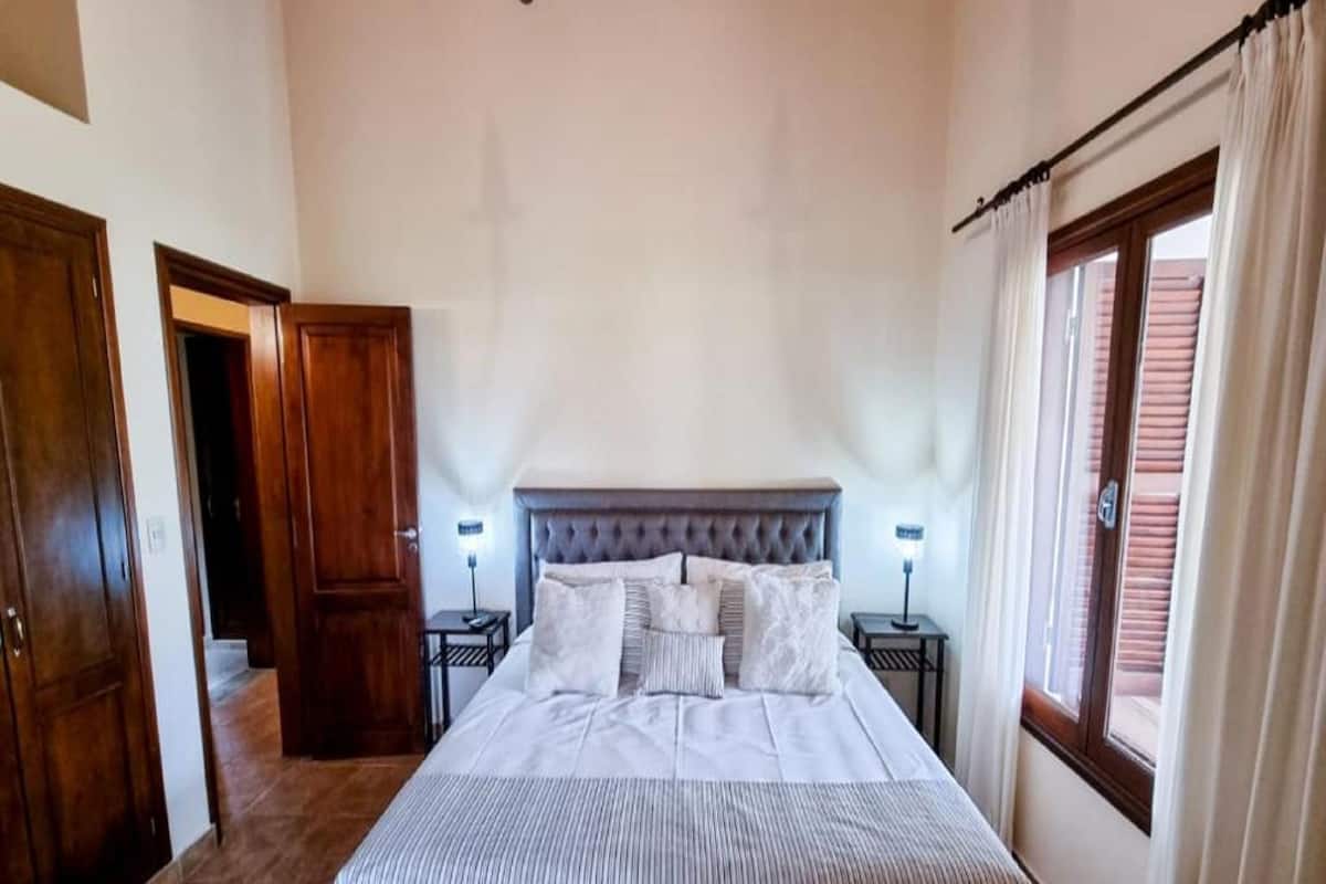 Ein Schlafzimmer mit einem großen Bett, zwei Nachttischen mit Lampen, einer Chandelier und einem Fenster mit Vorhängen.