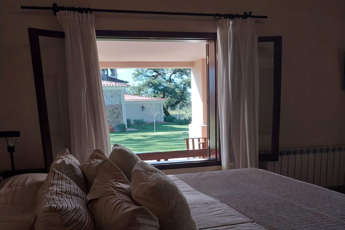 Ein Schlafzimmer mit einem großen Bett, weißen Vorhängen und Blick auf ein Gebäude und einen Rasen.