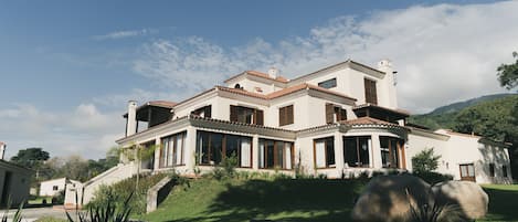 A large, two-story house with a tiled roof, multiple windows, and a stone wall in the foreground.