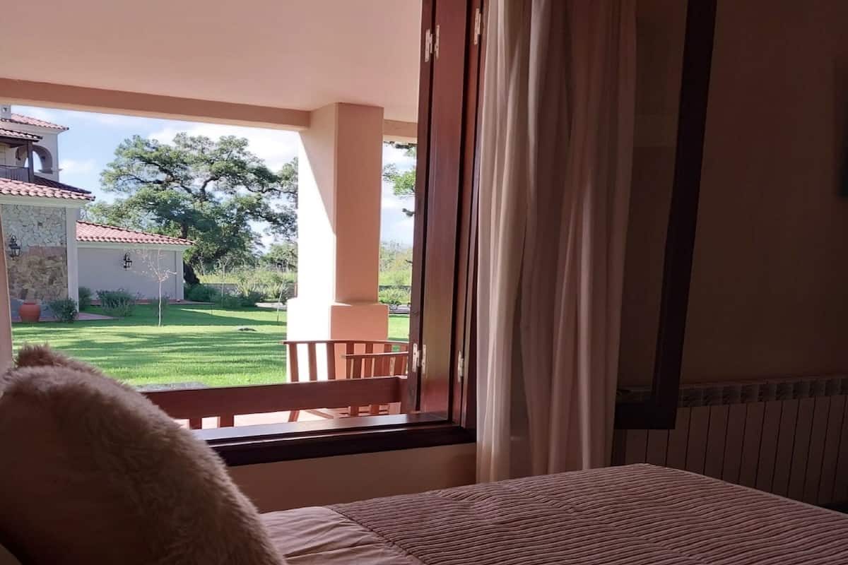 Ein Schlafzimmer mit einem großen Bett, einem Fenster mit Vorhängen und Blick auf einen Garten und ein Gebäude.